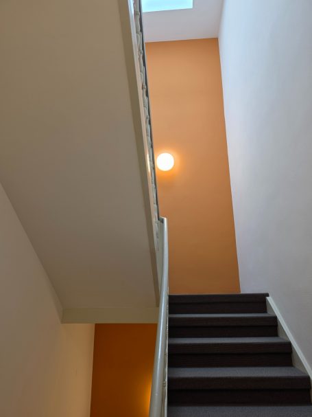 Treppenhaussanierung - Erneuerung der Decken- Wand- und Bodenflächen Treppenaufgang mit oranger Wand und einer Deckenlampe im oberen Bereich.